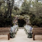 Beautiful outdoor ceremony set up at Ibiza Wedding Venue Ca Na Xica. A view up the aisle, wooden benches either side of the aisle lined with vibrant blue and purple flowers leading to a square floral arch