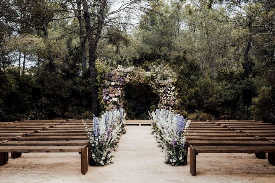 Beautiful outdoor ceremony set up at Ibiza Wedding Venue Ca Na Xica. A view up the aisle, wooden benches either side of the aisle lined with vibrant blue and purple flowers leading to a square floral arch