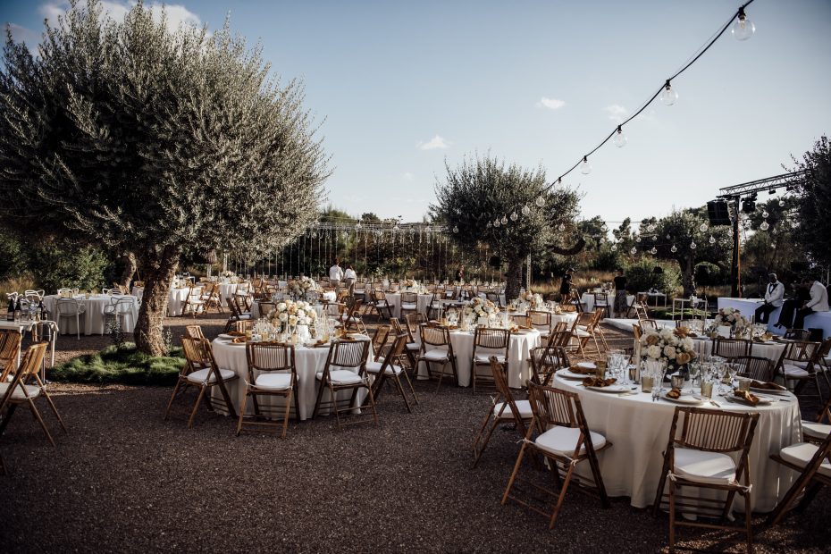 wedding breakfast tables set up ready for guests between olive trees and underneath fair lights at wedding venue ca na xica in ibiza