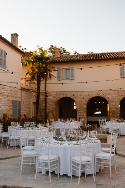 Round wedding tables with white wooden chairs and white linen tablecloths in a rustic courtyard