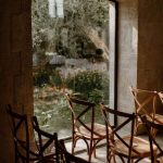 close up of wooden chairs set up for ceremony in renovated barn alongside arched window looking out over grounds at wedding venue in France