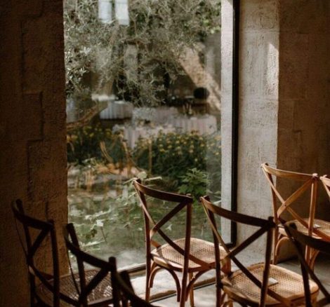 close up of wooden chairs set up for ceremony in renovated barn alongside arched window looking out over grounds at wedding venue in France