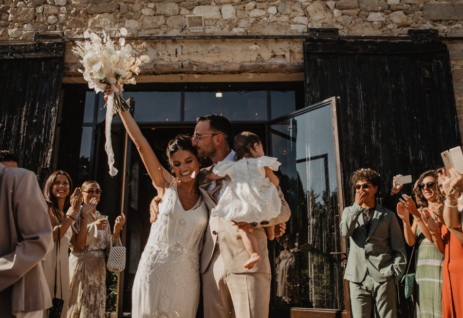 bride and groom holding baby whilst celebrating as coming out from ceremony at le Petit Roulet wedding venue in luberon in france