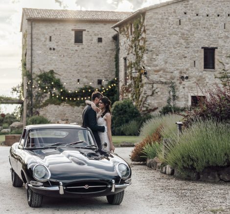 bride and groom lean on old black classic wedding car outside rustic wedding venue in umbria